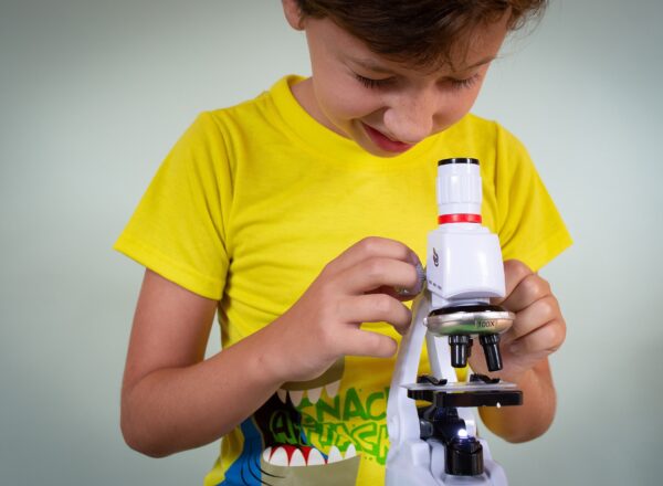 Kind mit gelbem T-Shirt steht vor einem Mikroskop und sieht durch das Okular.