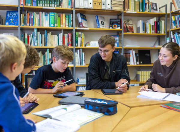 Eine Schülergruppe sitzt in der Bibliothek zu einer Gruppenarbeit mit Tablets und Heften